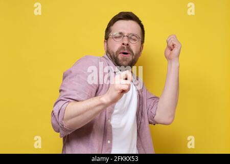 Homme caucasien de bonne humeur, il danse en faisant des mouvements avec les mains et chante avec les yeux fermés. Arrière-plan jaune. Banque D'Images