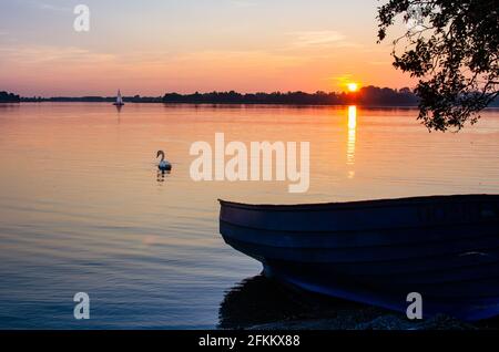 Lac Masurien, coucher de soleil, bateau à moteur sur la rive du lac. Au loin un cygne et un voilier Banque D'Images