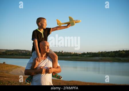 Le père tient son fils sur ses épaules sur fond de nature près du lac au coucher du soleil. Le garçon tient l'avion dans sa main. Concept de vacances en famille Banque D'Images