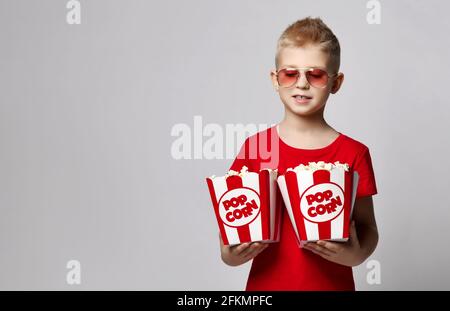 Petit garçon élégant en t-shirt rouge et porte-lunettes de soleil tenant deux boîtes de pop-corn entre les mains, prêt pour les rencontres au cinéma Banque D'Images