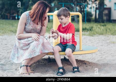 Maman et son fils sur l'aire de jeux. Garçon en T-shirt rouge vif et jeune femme en robe à l'extérieur l'été. La vie quotidienne. Concept de l'enfance et de la relation de Banque D'Images