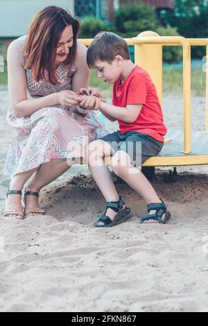 Maman et son fils sur l'aire de jeux. Garçon en T-shirt rouge vif et jeune femme en robe à l'extérieur l'été. La vie quotidienne. Concept de l'enfance et de la relation de Banque D'Images