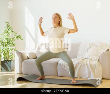 Femme active et mûre dans les vêtements de sport debout dans l'asana de yoga ou la posture du corps et regardant droit devant, s'exerçant à la maison dans le salon avec le soleil brillant à travers la fenêtre. Un mode de vie sain à la retraite Banque D'Images