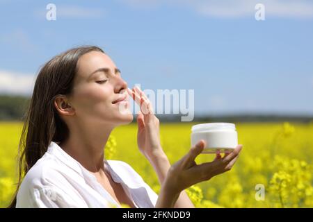 Femme de beauté tenant le pot appliquant la crème hydratante sur le visage dans un champ fleuri Banque D'Images