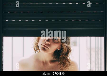 jeune femme aux yeux bleus et aux cheveux courts à l'intérieur de sa maison, assise à côté de l'aveugle fermé où son corps se reflète dans la fenêtre Banque D'Images