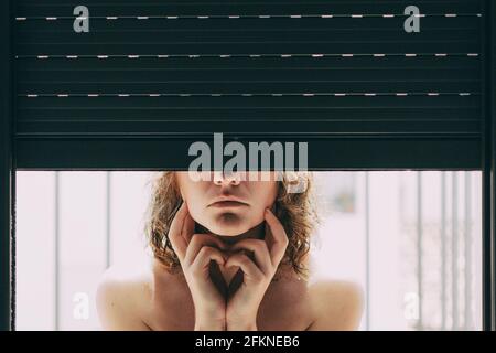 jeune femme aux yeux bleus et aux cheveux courts à l'intérieur de sa maison, assise à côté de l'aveugle fermé où son corps se reflète dans la fenêtre Banque D'Images