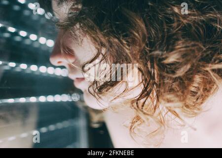 jeune femme aux yeux bleus et aux cheveux courts à l'intérieur de sa maison, assise à côté de l'aveugle fermé où son corps se reflète dans la fenêtre Banque D'Images