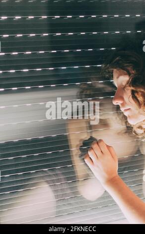 jeune femme aux yeux bleus et aux cheveux courts à l'intérieur de sa maison, assise à côté de l'aveugle fermé où son corps se reflète dans la fenêtre Banque D'Images