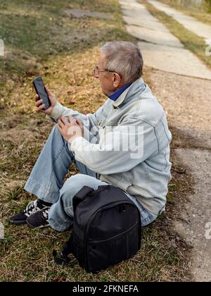 Un homme âgé en veste denim se trouve sur le côté de la route et regarde son smartphone. Sac à dos de voyage à proximité. Banque D'Images