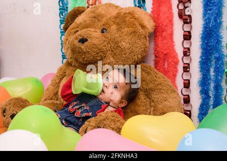 Petite petite fille indienne mignonne boire de l'eau penchée sur l'ours en peluche entouré de ballons, Mumbai Banque D'Images