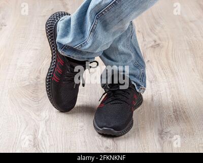 Homme debout à pattes croisées sur un sol brun clair. Homme en jeans bleus et baskets noires. Pieds debout dans des chaussures en mesh décontractées pour un style de vie actif. Banque D'Images