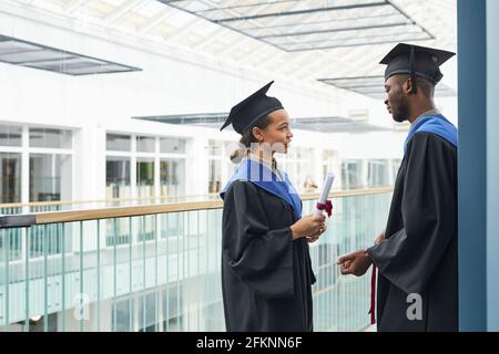 Vue latérale sur deux jeunes portant des robes de remise des diplômes bavardant à l'intérieur dans un intérieur moderne de l'université, espace de copie Banque D'Images