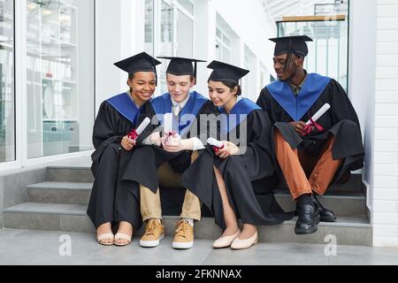Groupe diversifié de jeunes heureux portant des robes de remise des diplômes et utilisation d'un téléphone intelligent lorsque vous êtes assis sur des marches à l'extérieur Banque D'Images