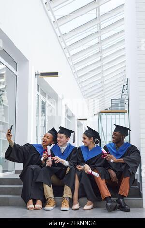 Vue sur toute la longueur de divers groupes de jeunes heureux porter des robes de remise des diplômes et prendre un selfie tout en étant assis sur les marches à l'extérieur Banque D'Images