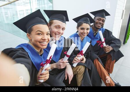 Vue en grand angle de divers groupes de jeunes heureux porter des robes de remise des diplômes et prendre le selfie à l'extérieur Banque D'Images