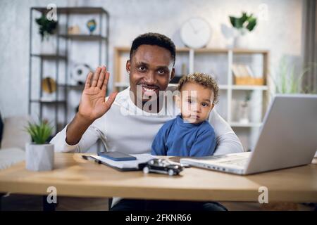 Homme africain souriant agitant sur l'appareil photo tout en étant assis au bureau à domicile avec son joli petit garçon sur les genoux. Concept de parentalité, de technologie et de freelance.