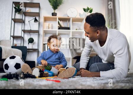 Jeune père afro-américain prenant soin de son fils mignon tout en restant ensemble à la maison. Homme heureux et bébé garçon dans des vêtements décontractés assis sur la moquette et jouant avec des jouets. Banque D'Images