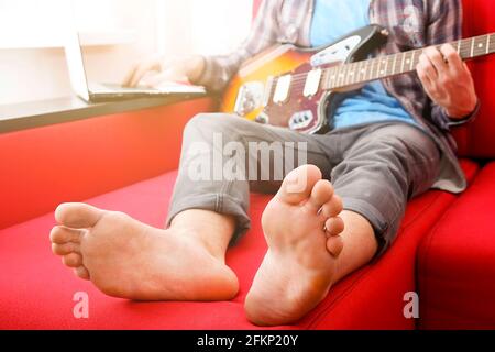 Homme pieds nus dans une chemise à carreaux, assis sur un canapé rouge près de la fenêtre jouant de la guitare électrique offset, cou en acajou. Rock guitariste joue de jazz impro Banque D'Images