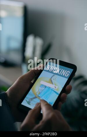 un jeune homme caucasien livre ses vacances en ligne avec son smartphone, avec un moteur de recherche simulé dans son écran, debout à son salon Banque D'Images