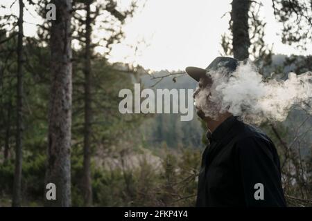 Cowboy fumant dans la forêt.Homme en chapeau noir et chemise sombre avec nuage de fumée au coucher du soleil.magnifique paysage boisé créatif avec espace de copie. Banque D'Images