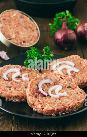 Boulettes de hamburger faites maison crues sur une assiette noire, une dans la presse à hamburger en arrière-plan, photo de stock verticale Banque D'Images