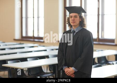 Portrait à la taille du jeune homme portant un peignoir de remise de diplômes et regardant la caméra tout en posant dans l'auditorium de l'école, espace de copie Banque D'Images