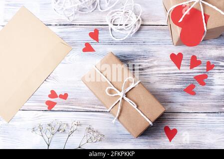 Cadeau dans un emballage en papier artisanal avec coeurs rouges en papier et enveloppe sur fond blanc en bois, emballage présent à la maison, vue de dessus, gros plan. Banque D'Images