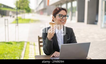 Femme souriante travaillant à distance sur un ordinateur portable et mangeant un cône de glace tout en étant assise à une table en bois à l'extérieur. Portrait d'une fille dans un costume d'affaires Banque D'Images