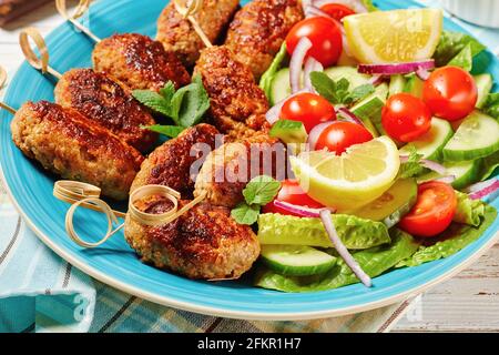 Kofta kebabs d'agneau haché, servi sur un plateau bleu avec sauce au yaourt, salade de légumes frais, et quartiers de citron, cuisine grecque sur un bois blanc Banque D'Images