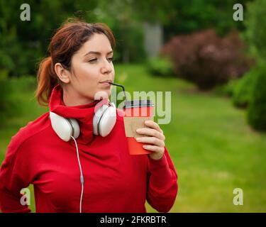 Femme aux cheveux rouges avec casque et boissons café avec une paille dans le parc. Une jeune fille aime se promener par une belle journée d'été et tient un cappuccino à emporter Banque D'Images