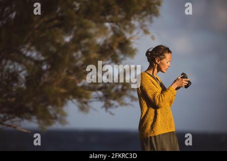 Jolie jeune femme avec un appareil photo sans miroir, prenant des photos au bord de la mer Banque D'Images