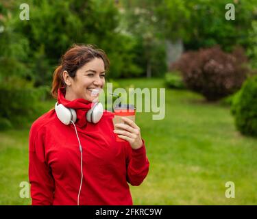 Femme aux cheveux rouges avec casque et boissons café avec une paille dans le parc. Une jeune fille aime se promener par une belle journée d'été et tient un cappuccino à emporter Banque D'Images