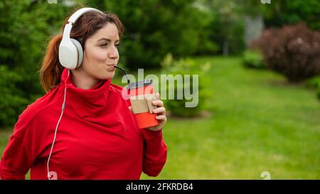 Une femme aux cheveux rouges écoute de la musique dans un casque et boit du café avec une paille dans le parc. Une fille aime une promenade sur une journée ensoleillée d'été et tient un Banque D'Images