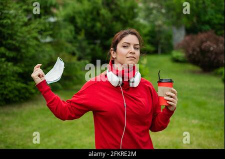 Une femme aux cheveux rouges dans un casque et boit du café avec une paille dans le parc et prend un masque médical. La fille bénéficie d'une promenade gratuite pendant un été ensoleillé Banque D'Images
