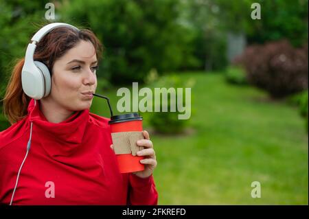 Une femme aux cheveux rouges écoute de la musique dans un casque et boit du café avec une paille dans le parc. Une fille aime une promenade sur une journée ensoleillée d'été et tient un Banque D'Images