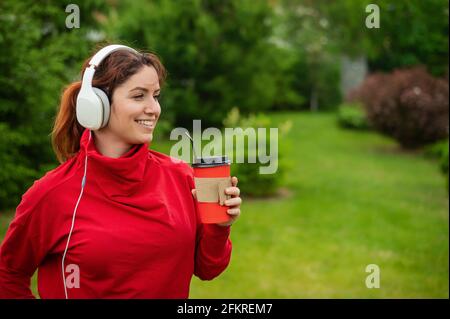 Une femme aux cheveux rouges écoute de la musique dans un casque et boit du café avec une paille dans le parc. Une fille aime une promenade sur une journée ensoleillée d'été et tient un Banque D'Images