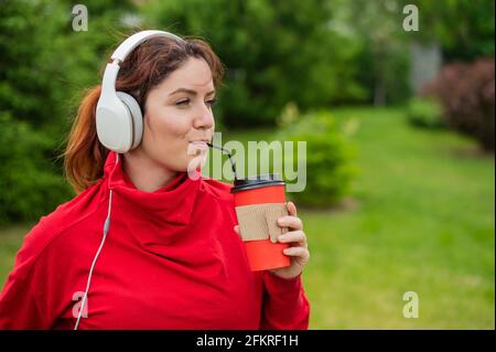 Une femme aux cheveux rouges écoute de la musique dans un casque et boit du café avec une paille dans le parc. Une fille aime une promenade sur une journée ensoleillée d'été et tient un Banque D'Images