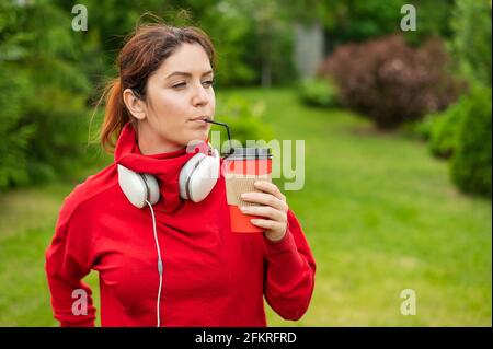 Femme aux cheveux rouges avec casque et boissons café avec une paille dans le parc. Une jeune fille aime se promener par une belle journée d'été et tient un cappuccino à emporter Banque D'Images