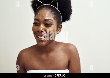 Belle jeune femme noire souriant sur fond blanc. Sur son visage est un masque hydratant doré. Concept de spa à la maison. Banque D'Images