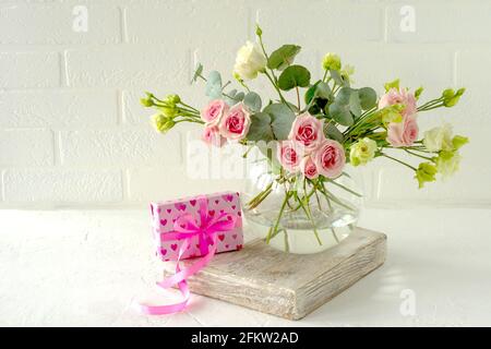 Bouquet de roses, d'euphorie et d'eucalyptus dans un vase en verre élégant sur une table à côté du cadeau d'amour. Composition des fleurs pour la décoration intérieure Banque D'Images