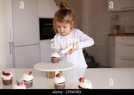 Mignon bébé fille 3-4 ans faire le cupcake, décorer avec de la crème fouettée sur la table de cuisine à la maison gros plan. Enfance. Banque D'Images