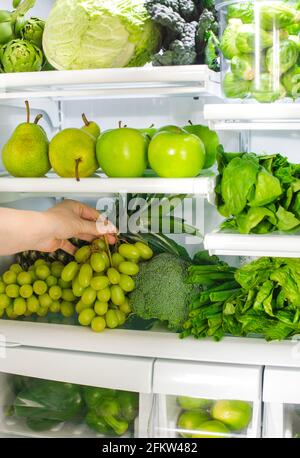 Fruits et légumes verts frais au réfrigérateur. La femme prend le bouquet de raisins verts du réfrigérateur ouvert. Une alimentation saine. Banque D'Images