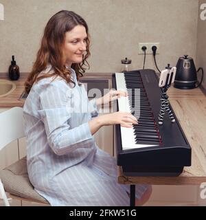 Une femme enceinte dans une queue de piano bleue joue le piano. Cours de musique dans la cuisine maison en gros plan Banque D'Images