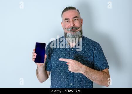 Grand boxeur blanc à barbe de 40-45 ans en chemise bleue regarder en ligne droite et afficher l'écran noir du smartphone tout en pointant à l'aide du Banque D'Images