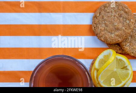 Biscuits aux flocons d'avoine, verre avec thé et tranches de citron, placés de façon décorative, vue du dessus sur une serviette rayée blanche et orange, avec espace libre pour Banque D'Images