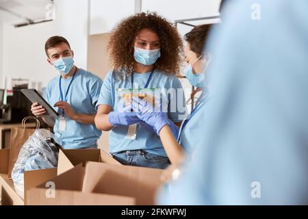 Des volontaires occupés portant un uniforme bleu, des masques de protection et des gants triant un paquet de pâtes et d'autres aliments tout en travaillant à des œuvres caritatives Banque D'Images