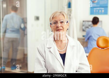 Gros plan d'une femme dentiste senior qui pose un diagnostic stomatologique à l'homme du patient pendant que l'infirmière médicale examine les prothèses à rayons X travaillant dans une armoire de stomatologie. L'équipe de l'hôpital rédige un traitement dentaire Banque D'Images