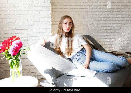 Jeune belle femme portant un t-shirt blanc sur un canapé en tissu gris à la maison. Jolie femme mince dans une situation domestique, reposant sur un canapé dans son haut Banque D'Images