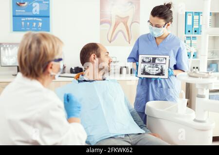 Malade assis sur un chaiar dentaire regardant la radiographie sur une tablette numérique discuter avec un médecin stomatologue senior sur le traitement de la douleur dentaire. Équipe de stomatologie médicale travaillant dans le bureau de la dentisterie Banque D'Images