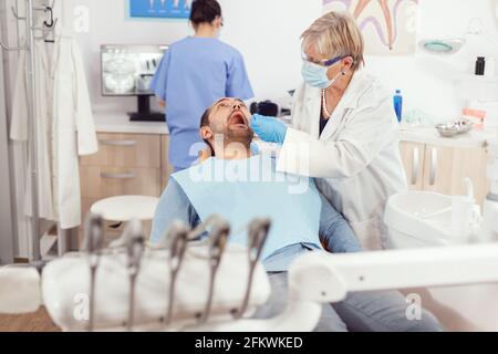 Dentisterie femme médecin senior faisant le nettoyage professionnel des dents à l'homme malade patient pendant la consultation orthodontique dans cabinet dentaire. L'équipe de l'hôpital examine les maux de dents qui préparent le traitement dentaire Banque D'Images